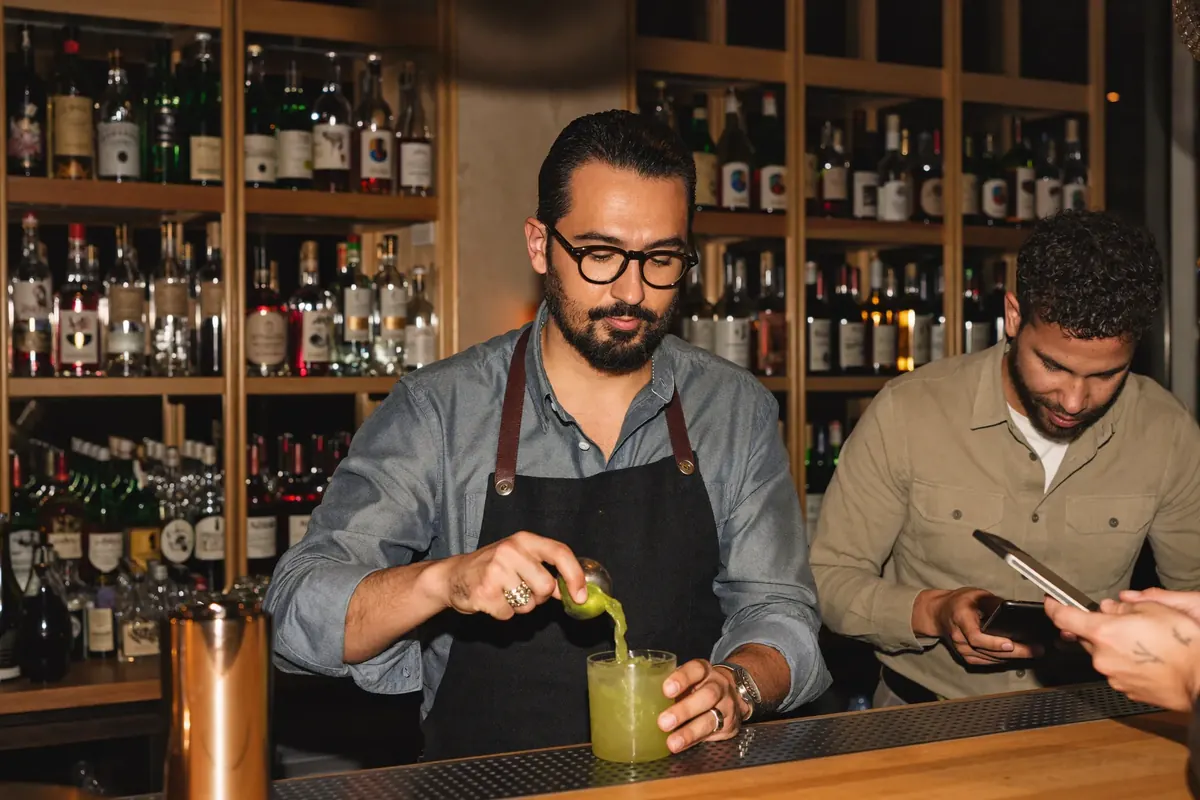 Bartender prepara un cóctel mientras un compañero revisa pedidos en una tablet